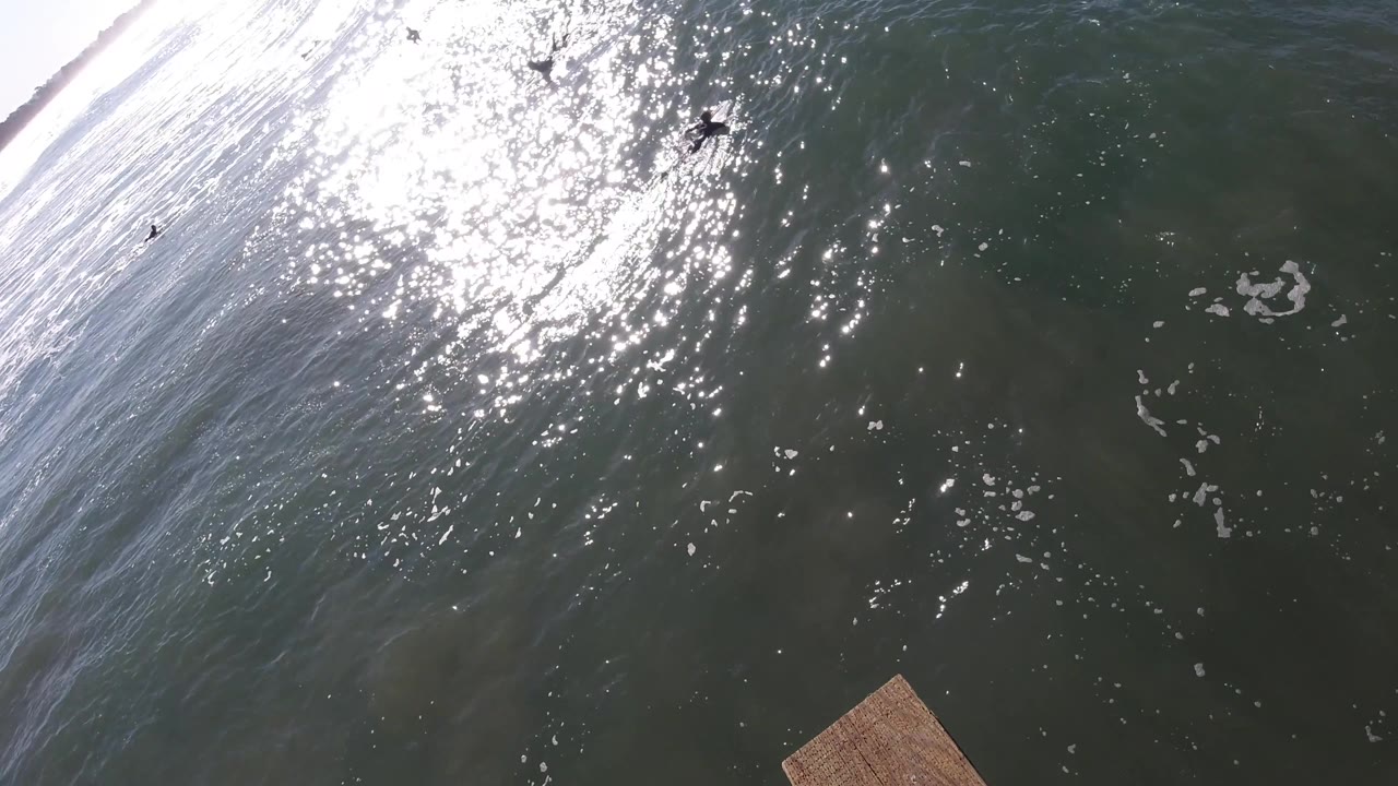 Pismo Beach surf is seen from above with strong sun sparkle on the dark water, a pale breaking wave along the left side, and no close visible people.