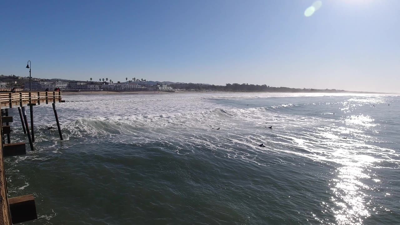 Wide Pismo Beach ocean view with the wooden pier edge on the left, bright sun glare on blue-green water, breaking white surf, distant shoreline, and small surfers visible offshore.