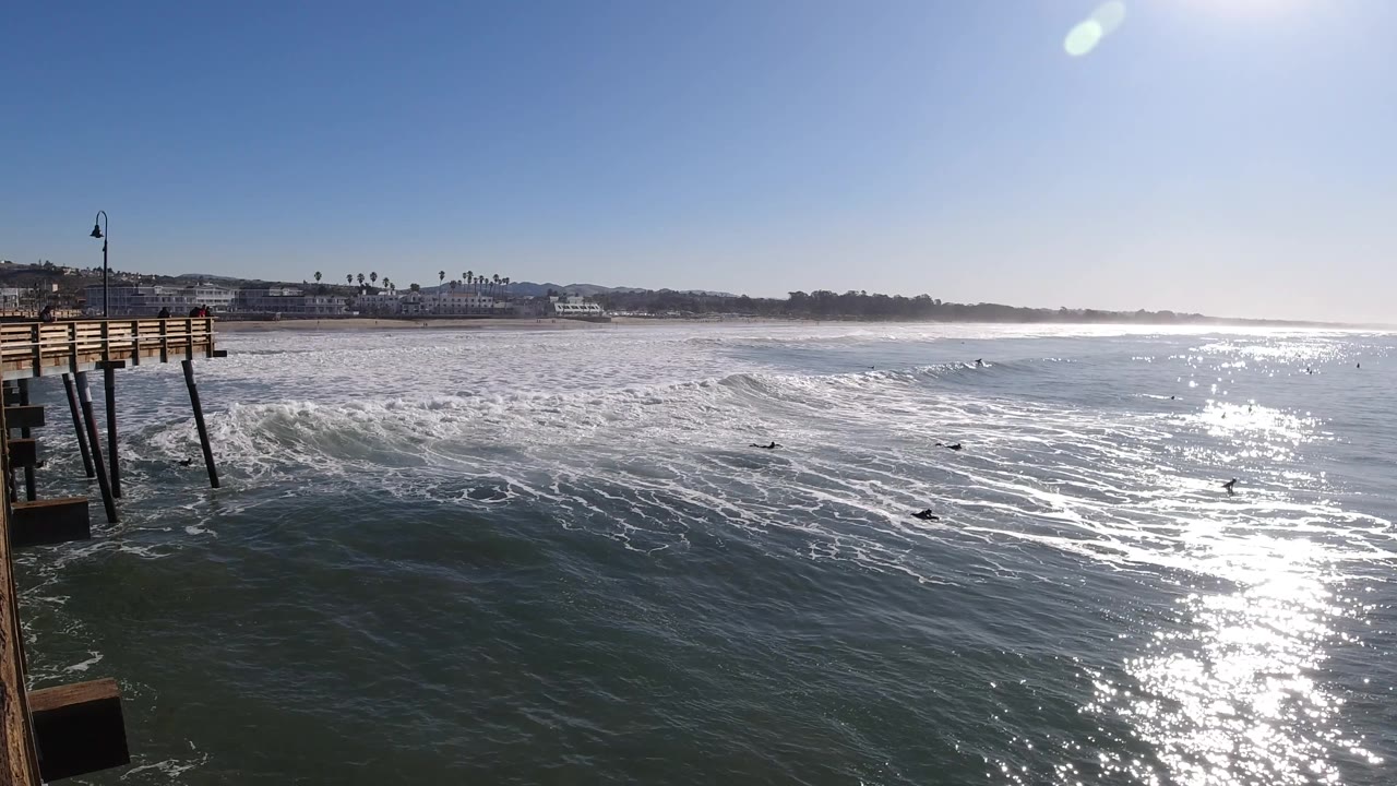 Wide Pismo Beach ocean frame with a surf line stretching across the middle, shimmering sun glare to the right, the distant shoreline in back, and small surfers scattered in the water.