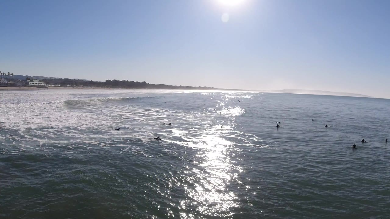 Wide aerial view of Pismo Beach ocean with bright sun glare on the water, small birds scattered offshore, surf breaking near the distant beach, and no visible people.