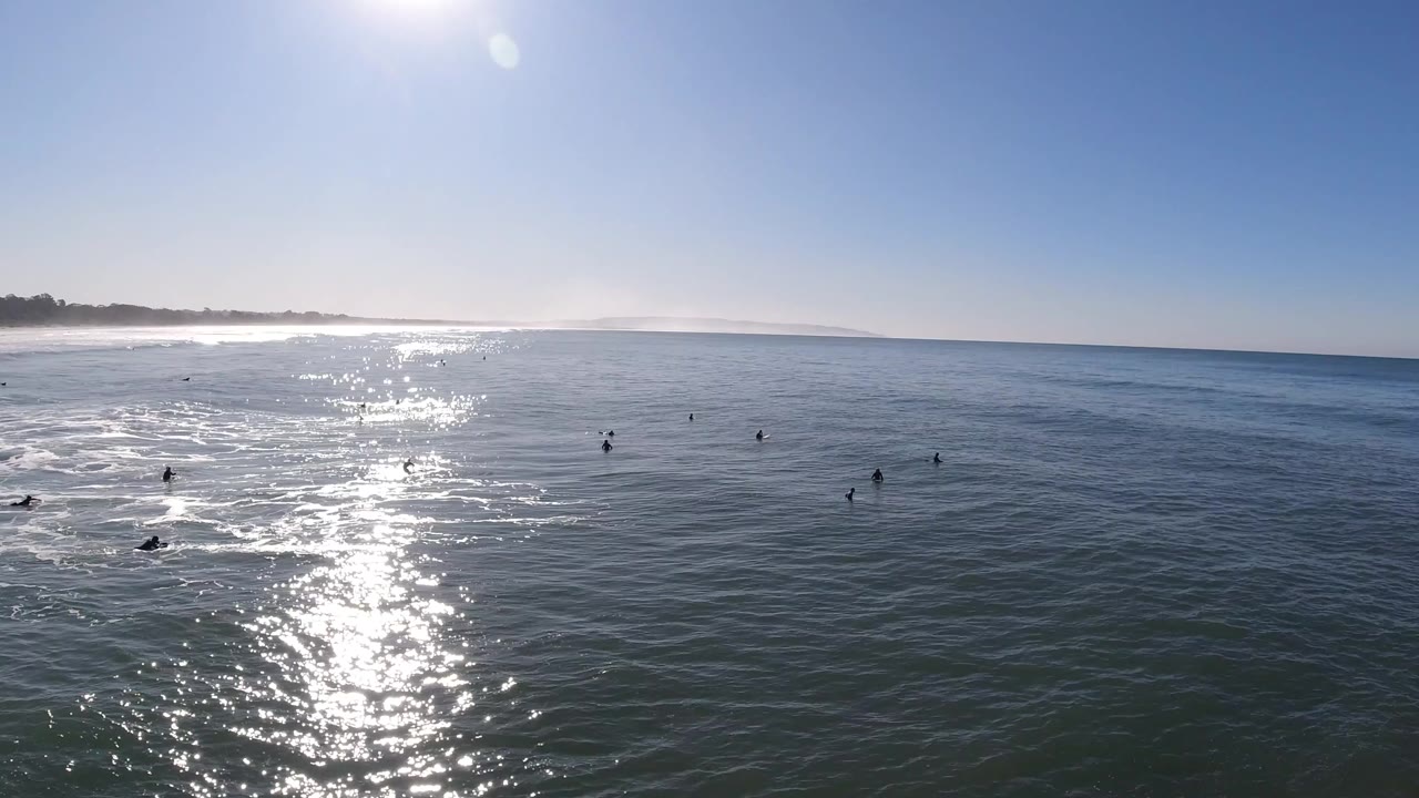 Surfers wait in open water under a bright sky with shoreline at left.