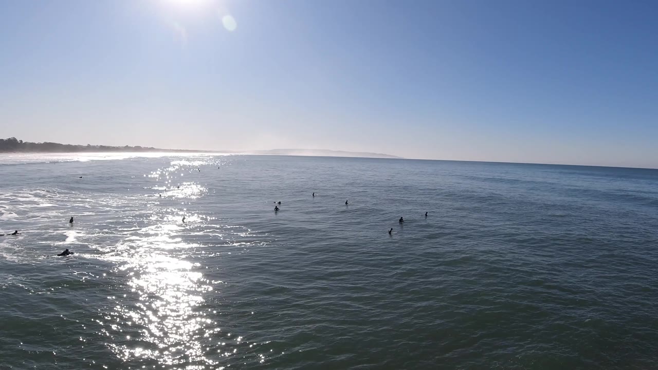 Pismo Beach aerial still with several distant surfers silhouetted against darker water to the right of a bright vertical sun reflection.