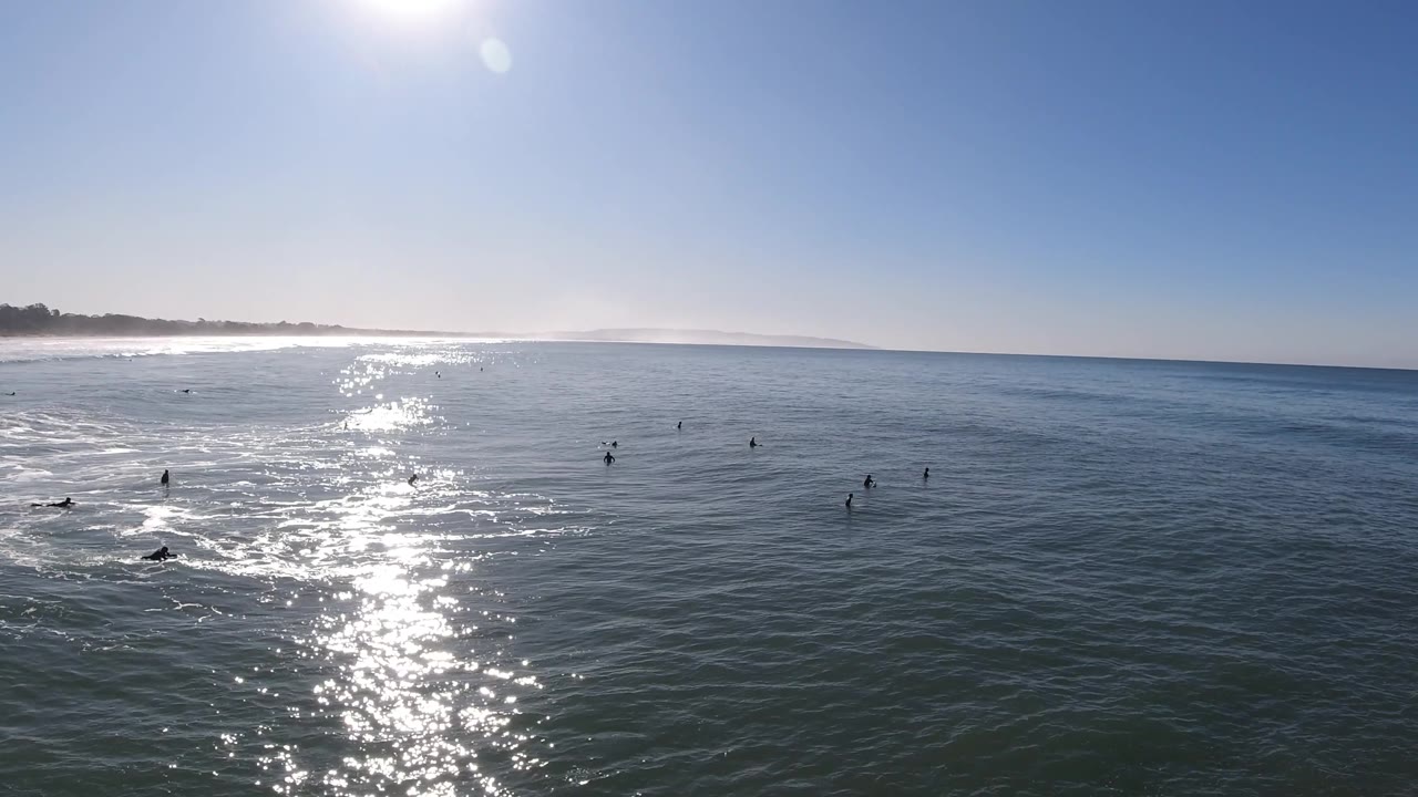 High ocean view with surfers near small wave foam and strong sun sparkle.