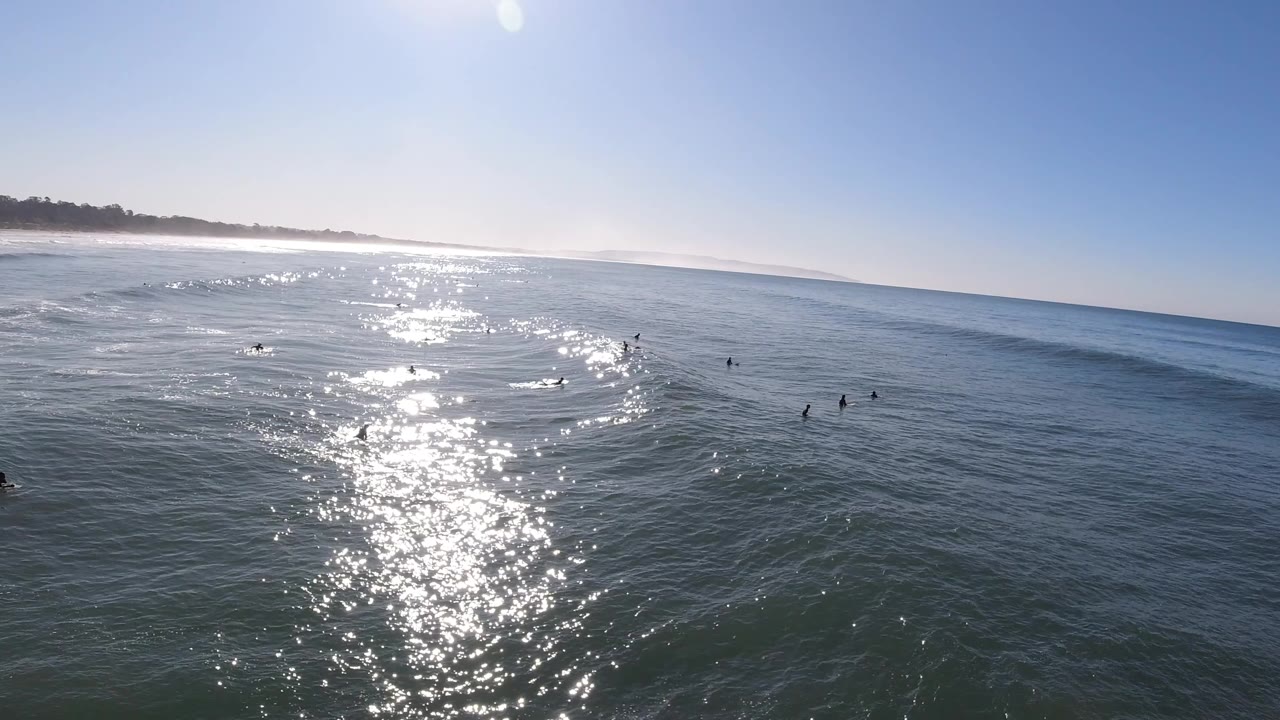 Aerial view over Pismo Beach ocean water with bright sun reflection, a distant shoreline at left, and small surfers or swimmers spread across the midground.