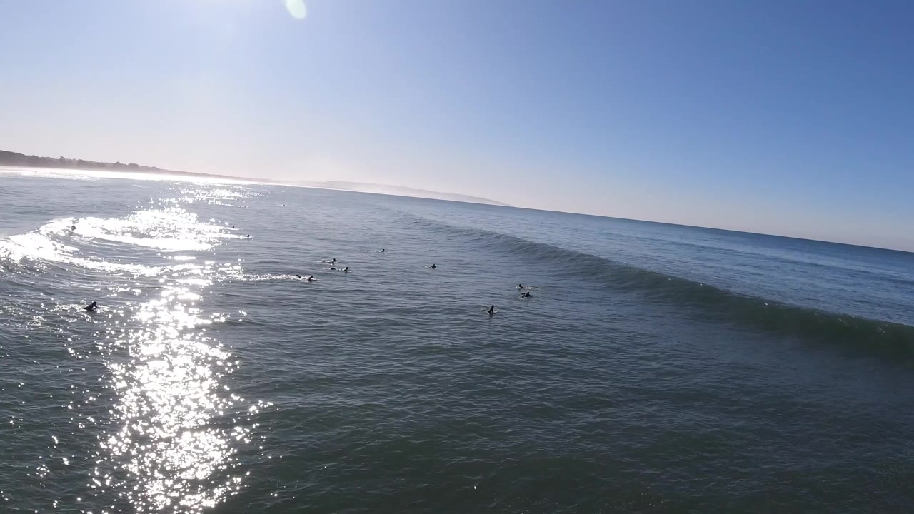 Wide aerial view at Pismo Beach with sun glare across the ocean, small surfers in the water, and a smooth swell beginning to rise.