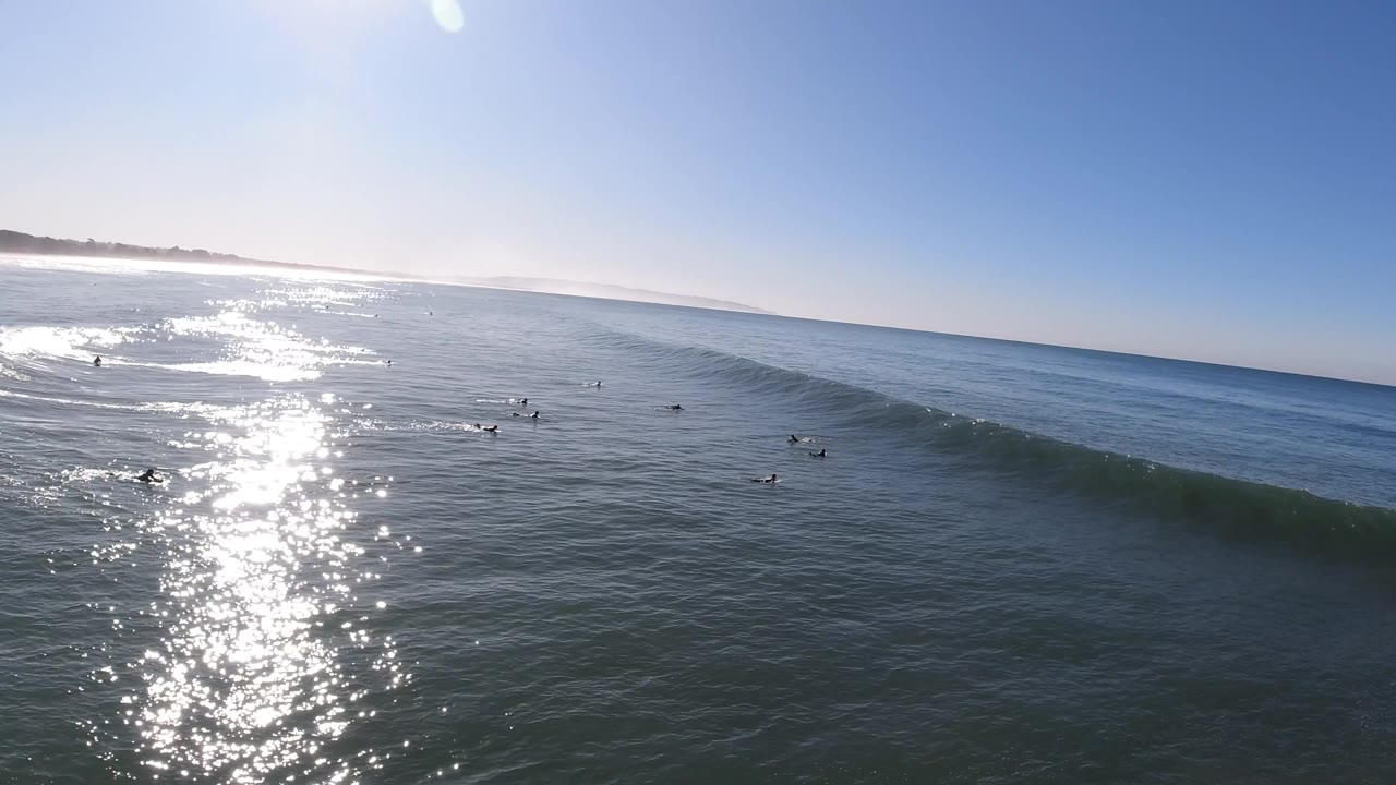 A wide Pismo Beach surf view shows the swell more defined, with small surfers scattered in the midground and bright sun shimmer stretching across the water.