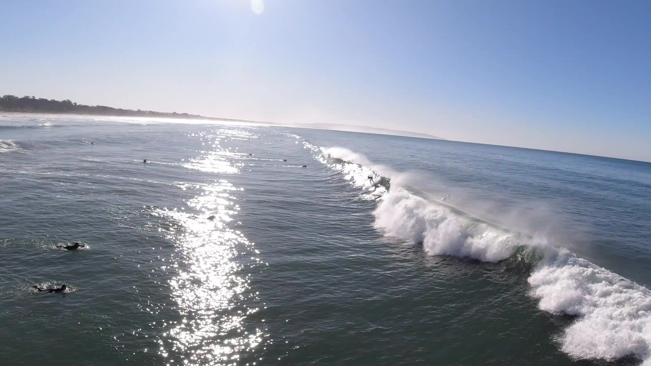 A high view over Pismo Beach water shows a large breaking wave crossing the frame, bright sun glare on the ocean, distant shoreline, and tiny surfers with no recognizable faces.