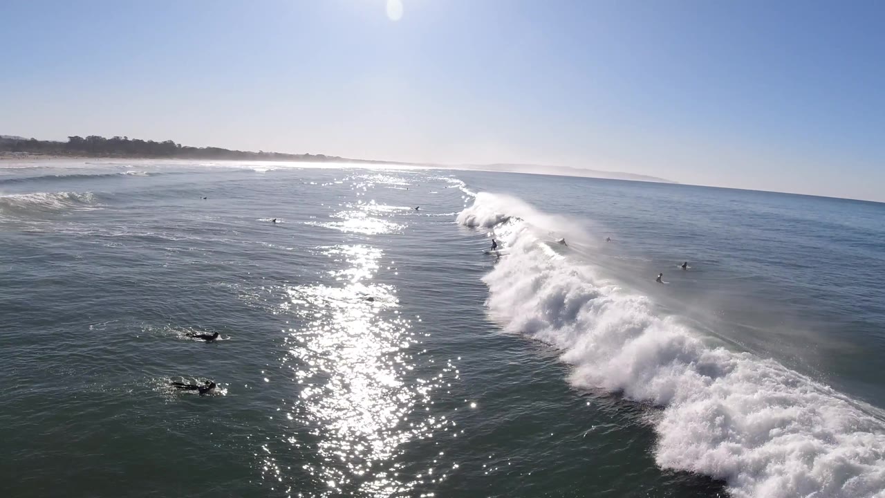 A broad white wave breaks near the center of the Pismo Beach frame, with dark small surfer shapes in the surrounding water, strong glare, and a low coastline on the horizon.