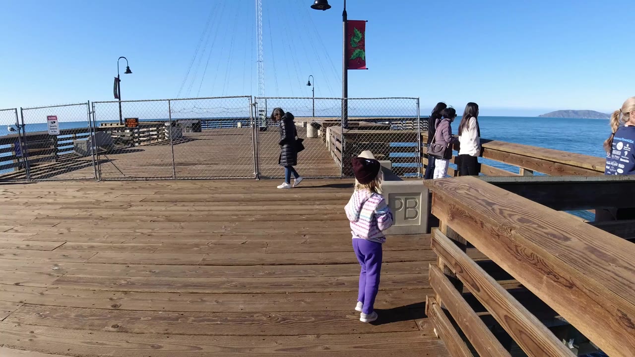 A child in purple pants stands on a wooden oceanfront platform in Pismo Beach while adults gather near the railing and the ocean sits behind fencing and benches.