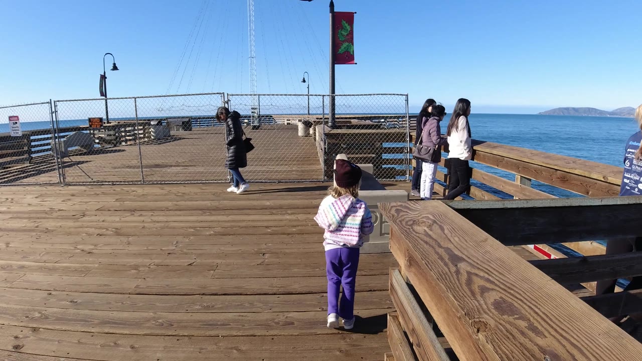 A child pauses near adults at the Pismo Beach pier railing, with the ocean, surf line, distant hills, fencing, and a bench visible in bright daylight.