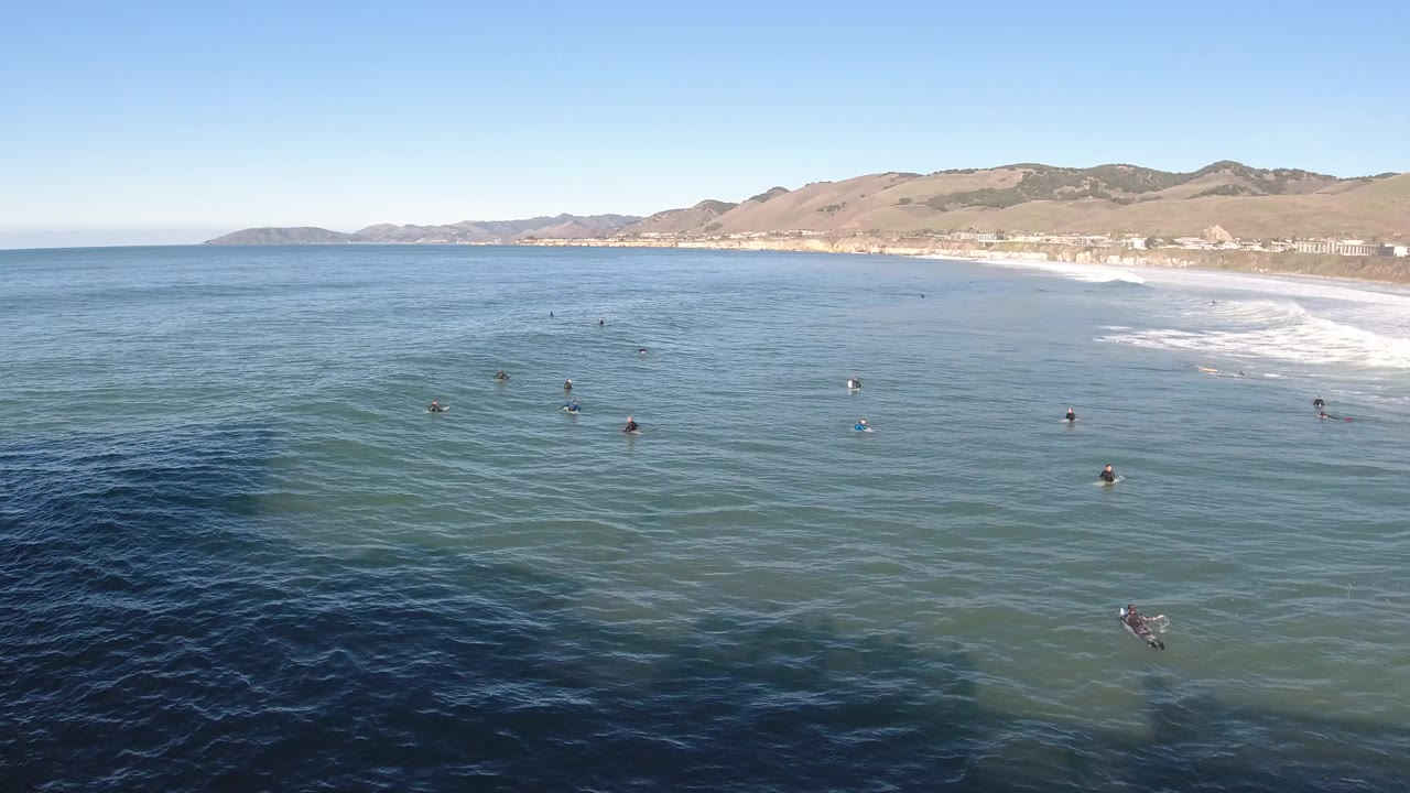Wide aerial view over Pismo Beach, California, with small swimmers scattered in blue-green ocean, white surf along the right shoreline, and low hills beyond the beach.