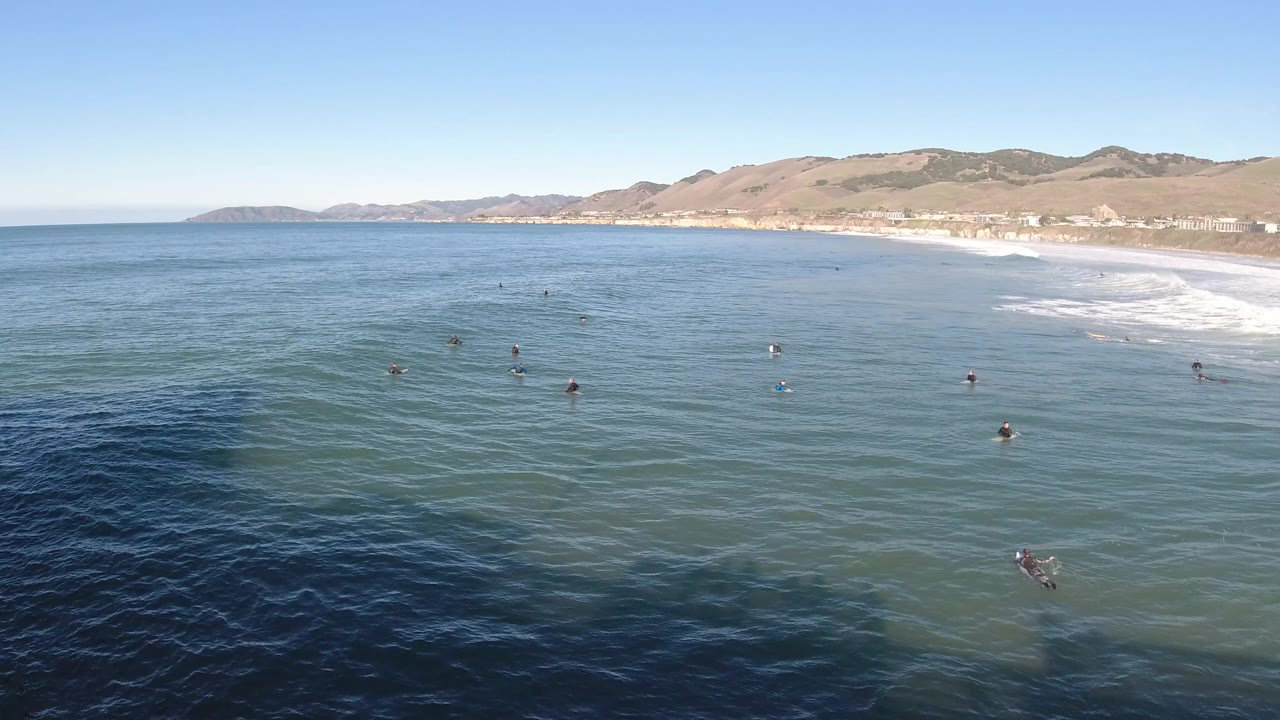 Aerial ocean view with swimmers spread across calm water and surf near the beach.
