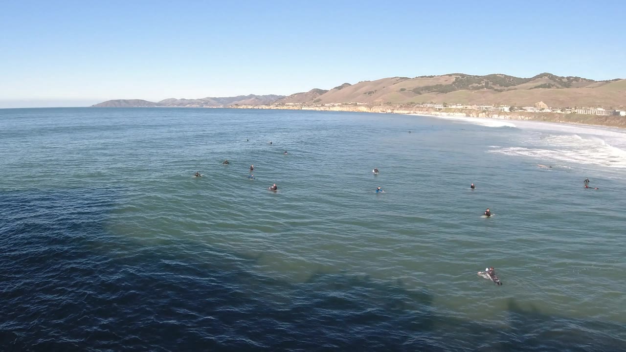 Wide elevated frame over Pismo Beach showing scattered swimmers and surfers in the blue water, with the shoreline and hillside community running across the background.