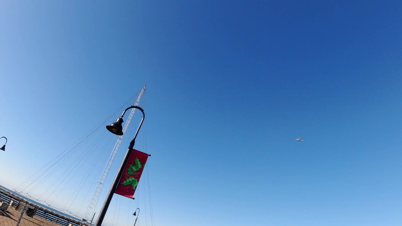 Opening frame in Pismo Beach, California, with a canted pier lamp and red banner against a clear blue sky, a sliver of waterfront railing below, and no visible people.