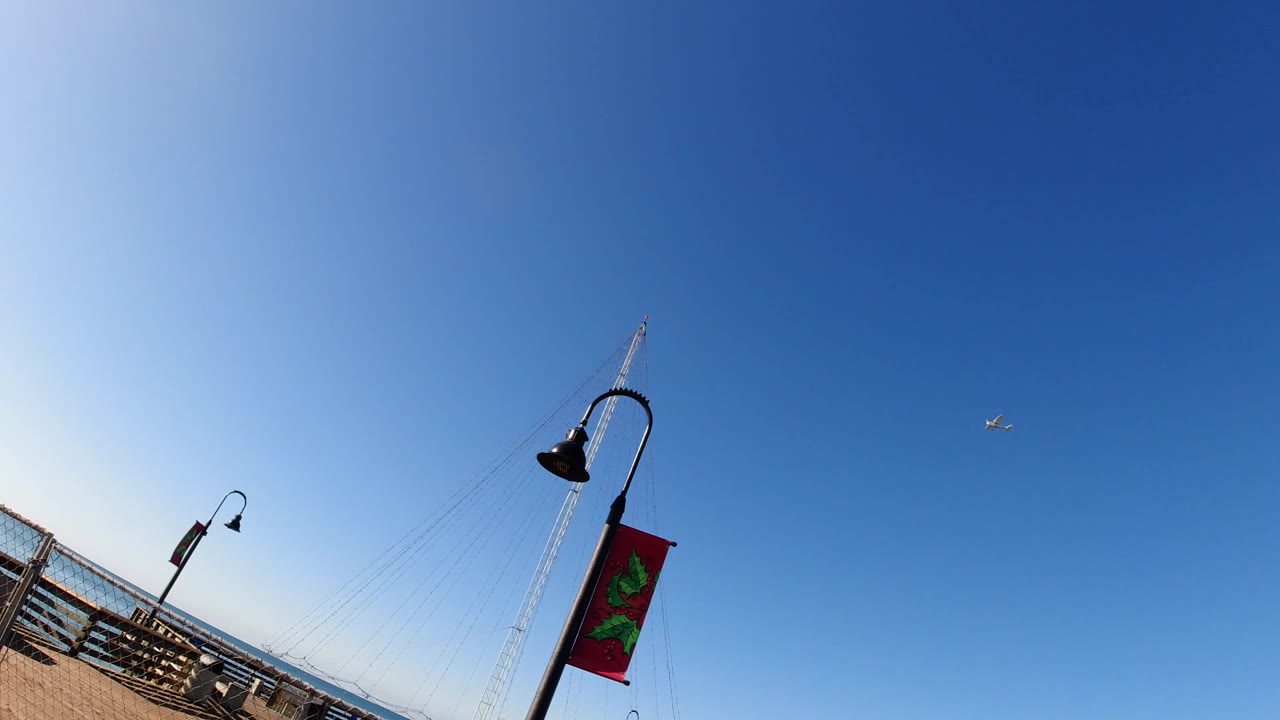 Third frame in Pismo Beach, California, shows more of the pier railing entering from the left, with the lamp post and banner set against a wide blue sky and no visible people.