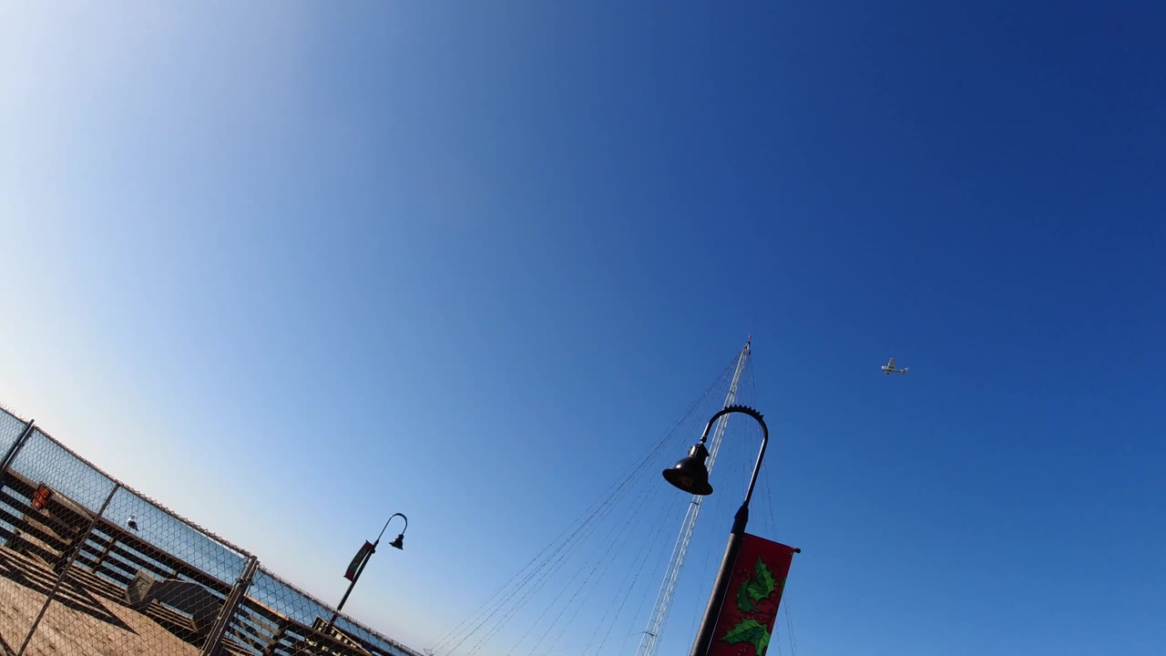 Fourth frame in Pismo Beach, California, places the pier railing and deck diagonally across the bottom left, with lamp posts, a small aircraft-like speck in the sky, and no visible people.