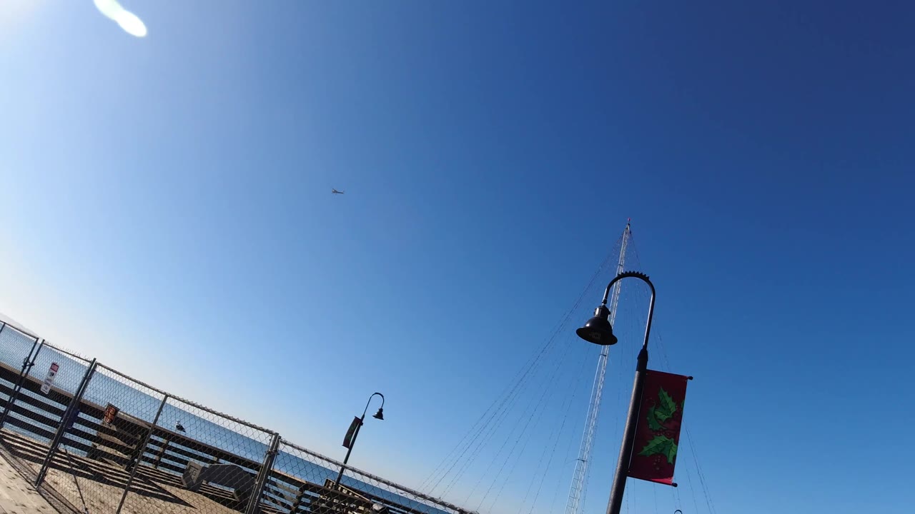 A canted Pismo Beach boardwalk frame keeps the lamp posts and red banner high in the image, with benches and fencing leading toward the ocean and no visible people.