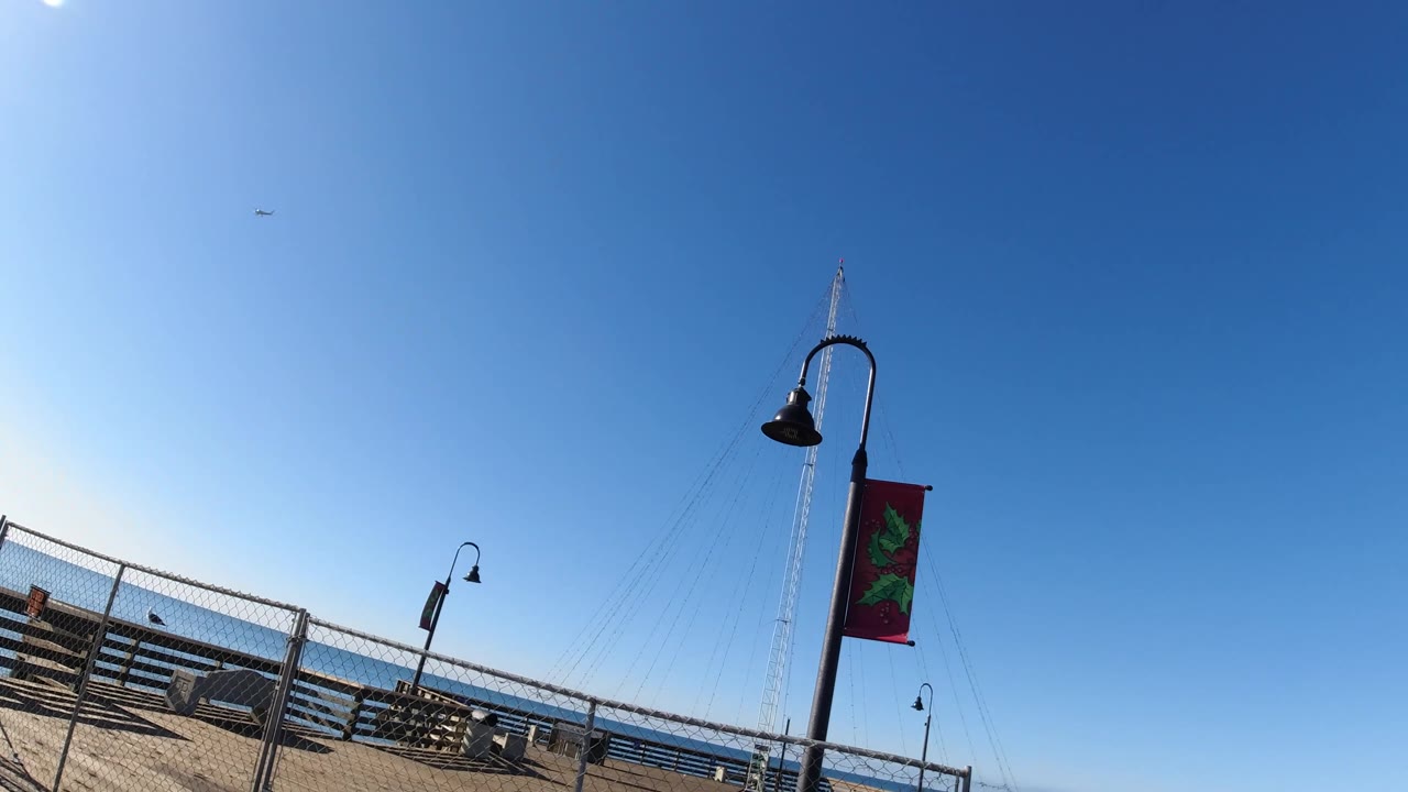 The Pismo Beach boardwalk appears at a steep angle with a tall lamp and red banner near the right side, smaller lamps along the fence, benches, ocean, and no visible people.