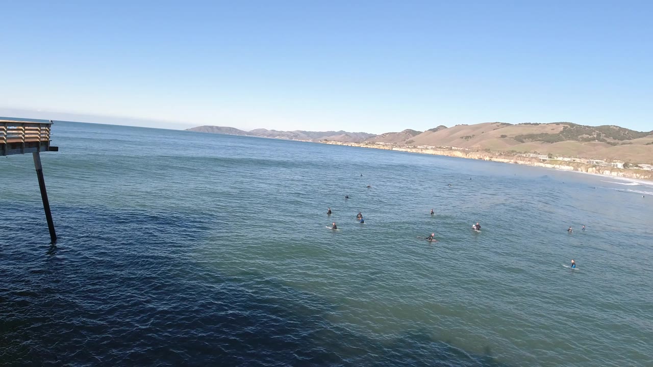 Pismo Beach ocean view with blue-green water, small people offshore, distant beach and hills, and the edge of a pier structure entering from the left.