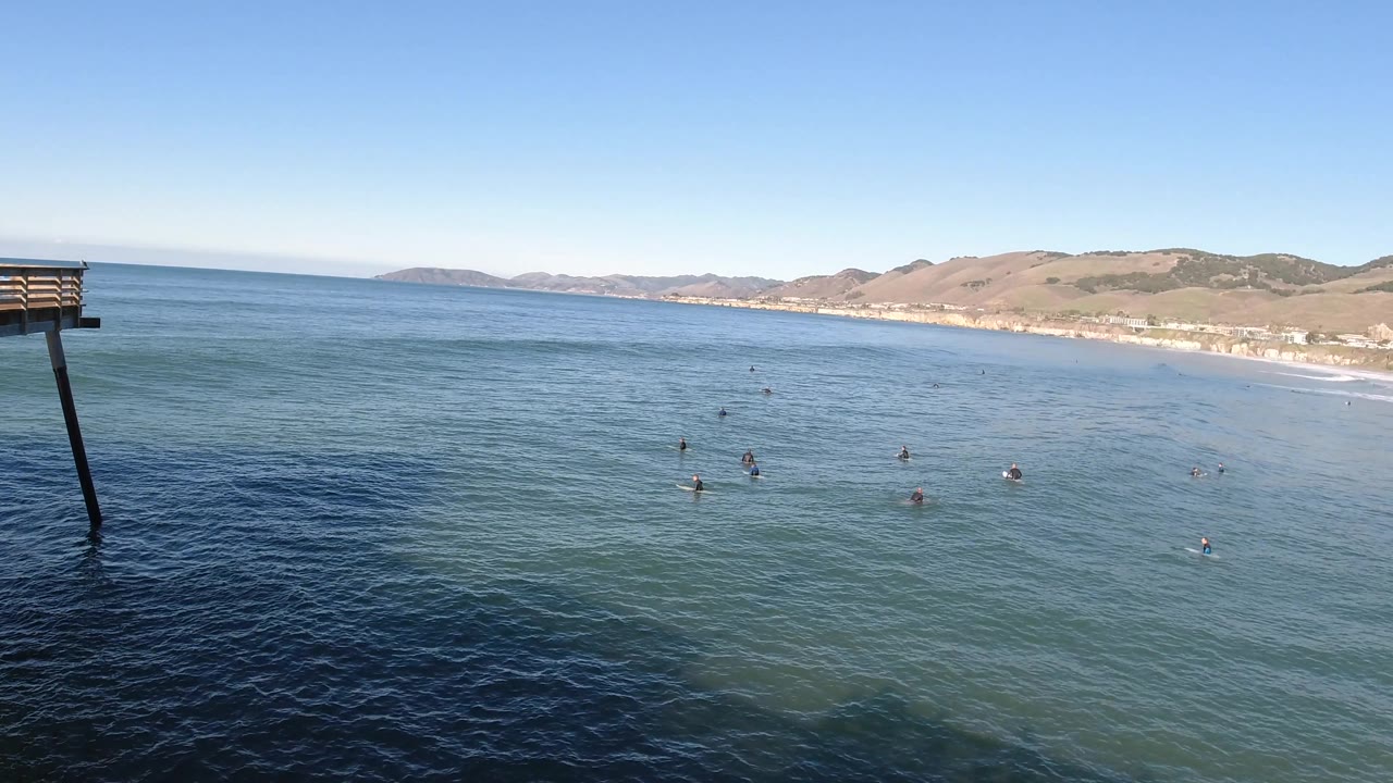 Pismo Beach ocean scene with a dark pier support dropping into the water, small people offshore, distant shoreline buildings, and hills under a clear sky.