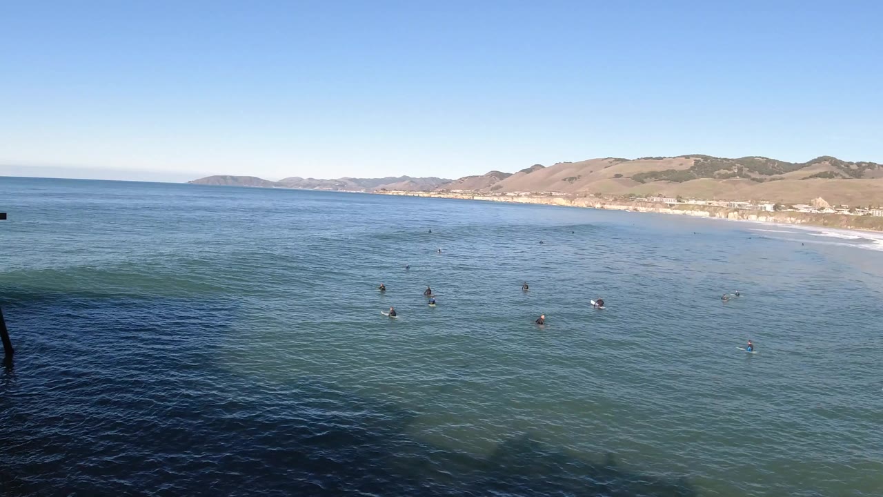 Wide elevated view over Pismo Beach, California, with small bathers spread across blue-green ocean water, a sandy shoreline at right, and low hills in the distance.