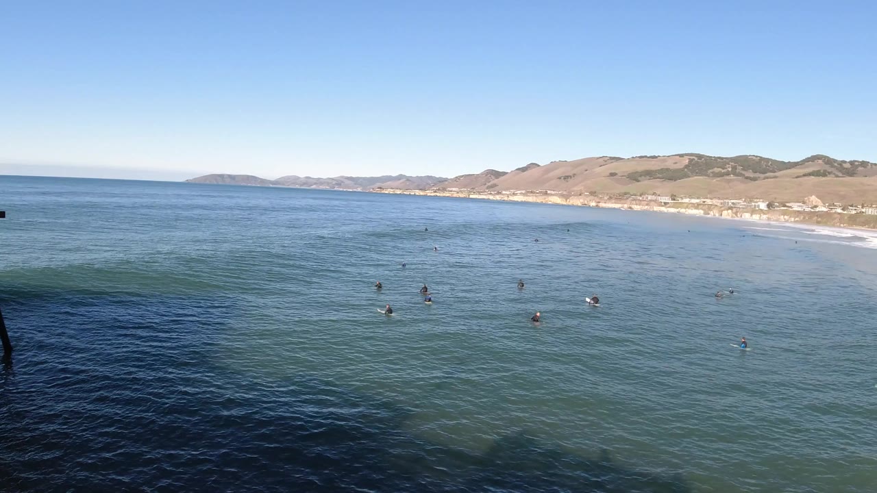 Pismo Beach water fills the foreground as scattered bathers remain offshore, with the surf line and hillside coast stretching across the background.