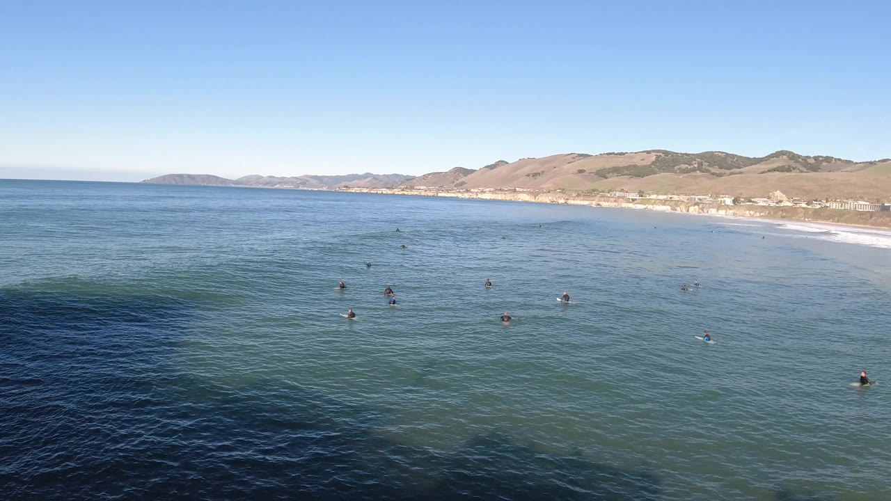 Elevated Pismo Beach view with bathers clustered near the center of blue water, a pale wave band near shore, and sunlit hills beyond the beach.