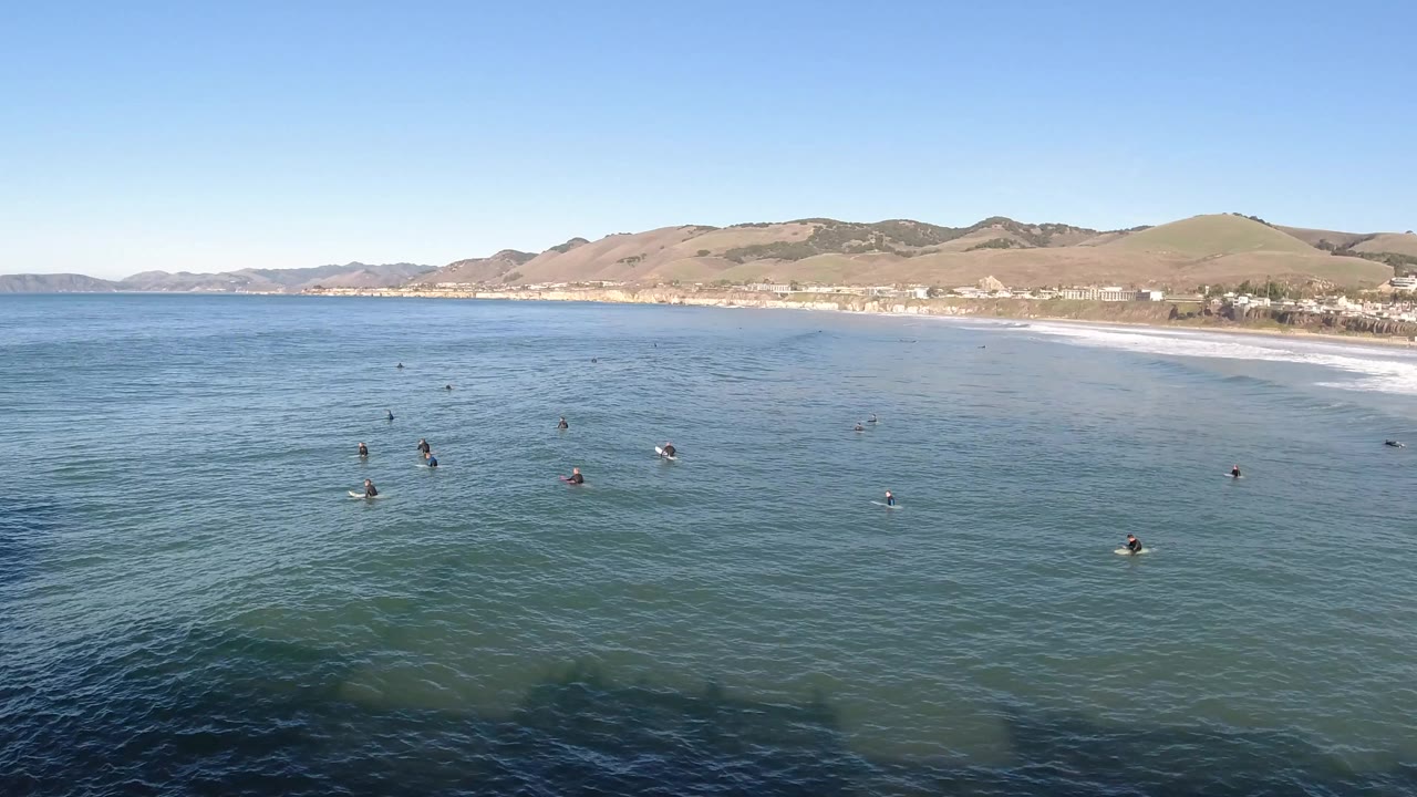 Aerial frame over Pismo Beach, California showing scattered people in blue-green nearshore water, white surf along the beach, tan hills, and a clear sky.
