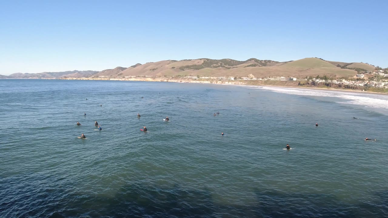 Pismo Beach, California appears from above with scattered swimmers or surfers in the water, white waves reaching the shore, hillside terrain, and beachfront structures in the distance.