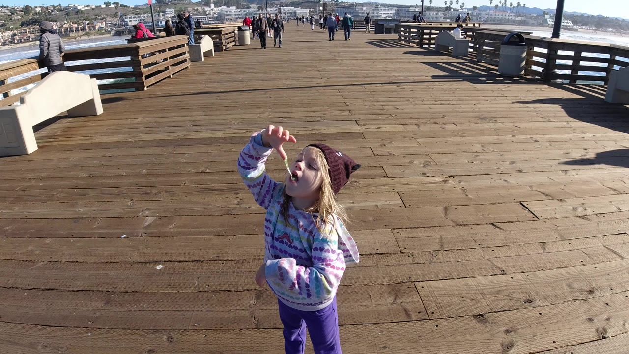 At Pismo Beach, California, the girl remains in the foreground eating, with long wood planks, a white bench, railings, and small groups of people spread across the pier in bright sun.