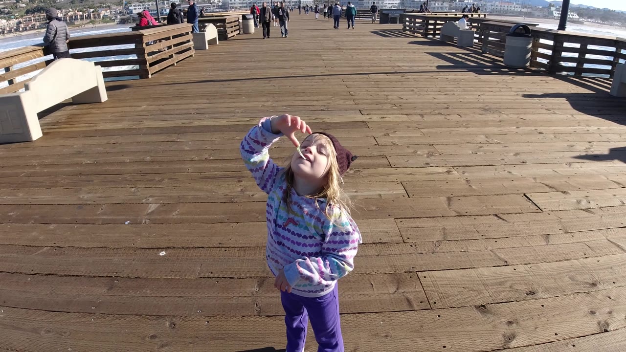 At Pismo Beach, California, the child looks upward with her hand near her face, standing on the wooden pier as pedestrians and railings recede into the background.