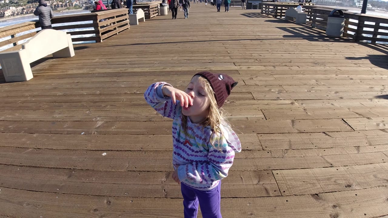 Young girl eating with the pier railing and bench behind her.