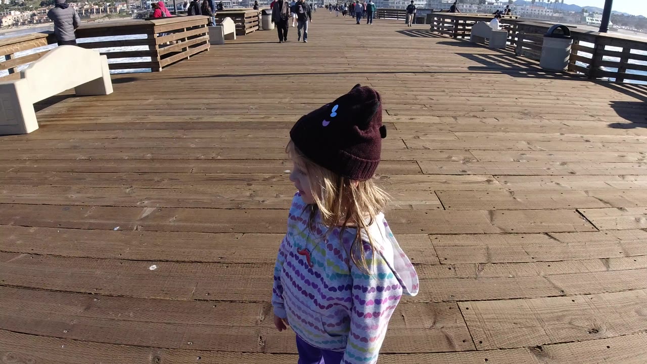 A young girl in a striped hoodie and dark knit hat stands near the lower left of the Pismo Beach pier as benches, railings, ocean, and distant pedestrians fill the background.