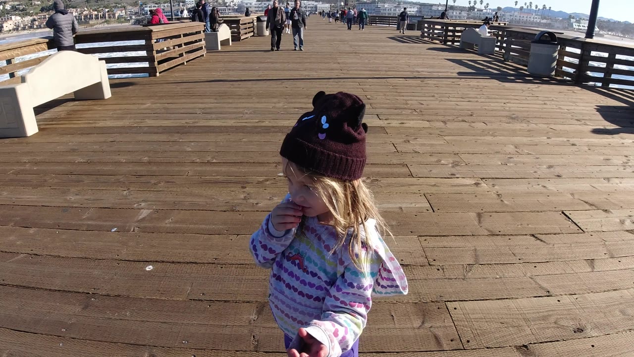 The girl turns toward the right side of the Pismo Beach pier frame, with sunlit planks, a bench, pier railing, ocean water, and distant people visible behind her.