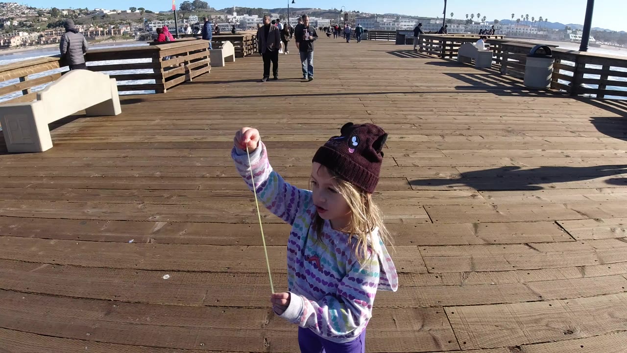 At Pismo Beach, a young girl in a maroon hat stretches gum from her mouth on the wooden pier while benches, pedestrians, ocean, and hillside homes sit behind her.