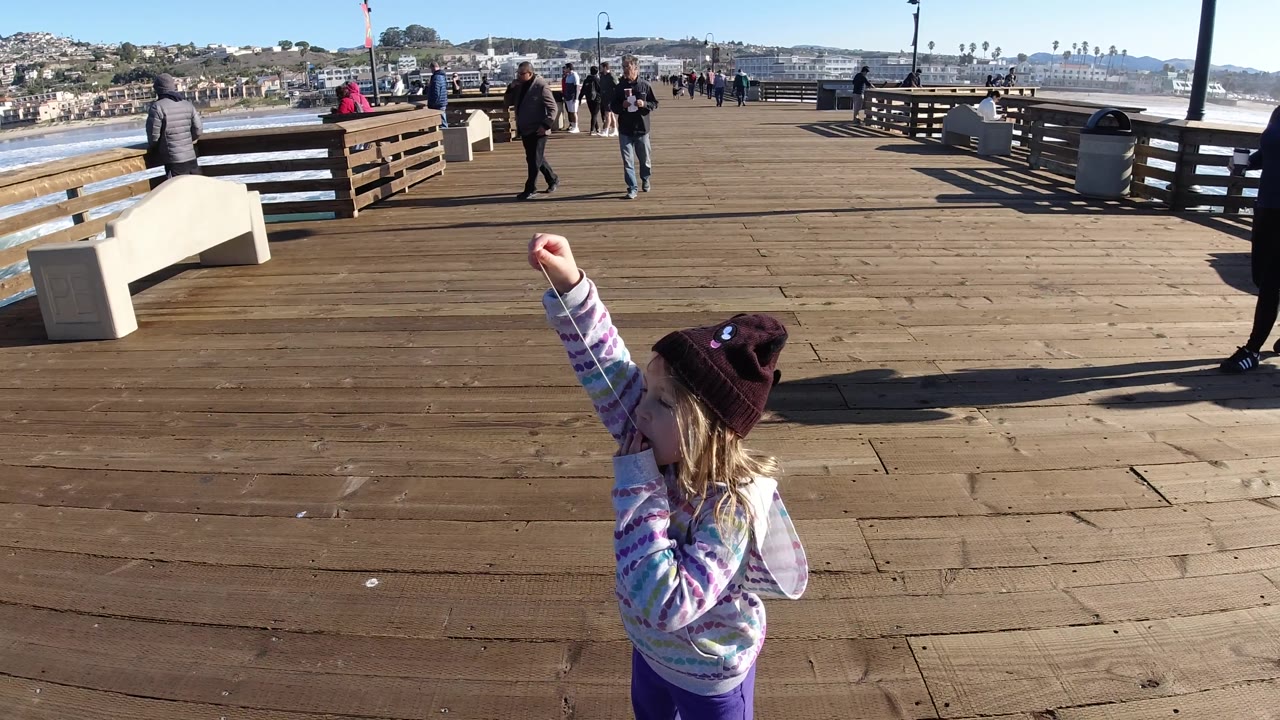 At Pismo Beach, the girl raises the gum toward her mouth near the lower right of frame, with open pier boards, benches, railings, pedestrians, ocean, and distant hills visible.