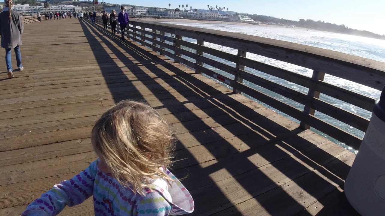 A blonde child appears close to the lower foreground on a wooden pier in Pismo Beach, California, with pedestrians ahead, ocean surf to the right, and long railing shadows across the boards.