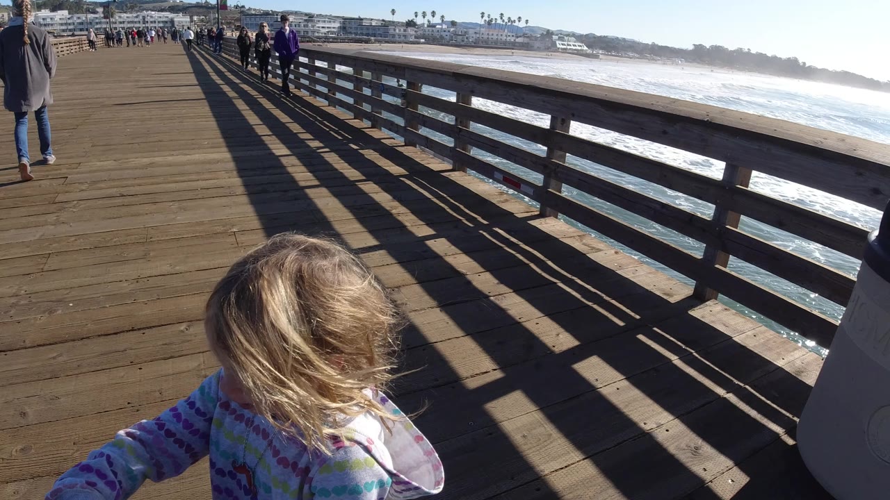 Blonde child at the lower edge of a sunny pier scene with people walking by the ocean.