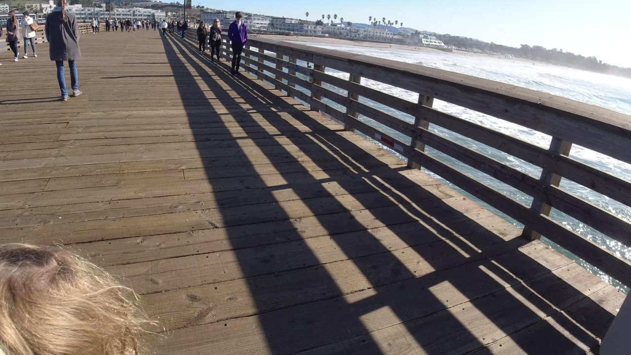 Only the top of the blonde child's head is visible at the bottom of the Pismo Beach pier view, while pedestrians move along the deck and sunlight throws dark railing shadows toward the ocean side.