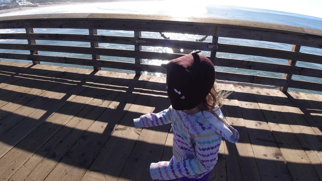 Young girl in a dark animal-eared hat walks along a wooden pier at Pismo Beach, California, with bright ocean glare beyond the railing.