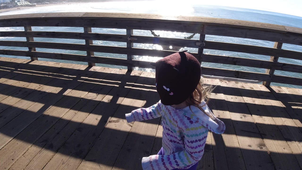 The child continues along the Pismo Beach pier, seen from above with long rail shadows crossing the wood planks and surf visible past the railing.