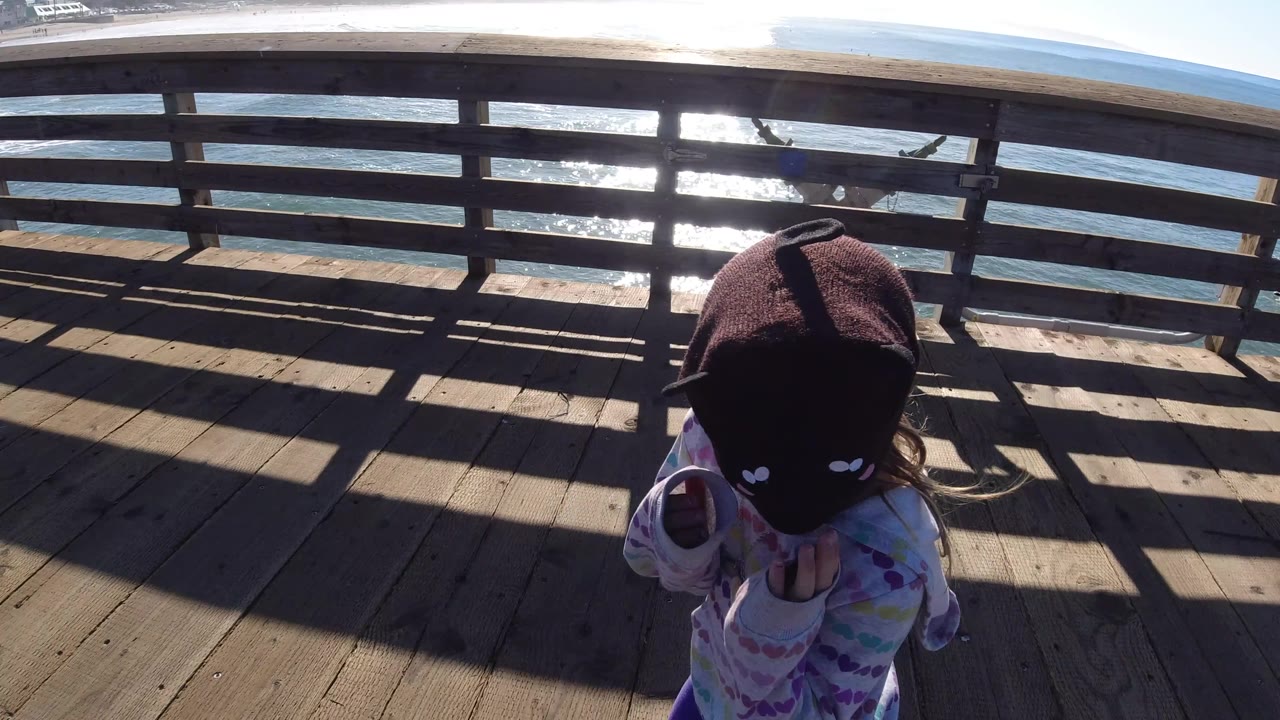At Pismo Beach, California, the girl moves toward the right edge of the frame while the pier railing and glittering ocean fill most of the view.