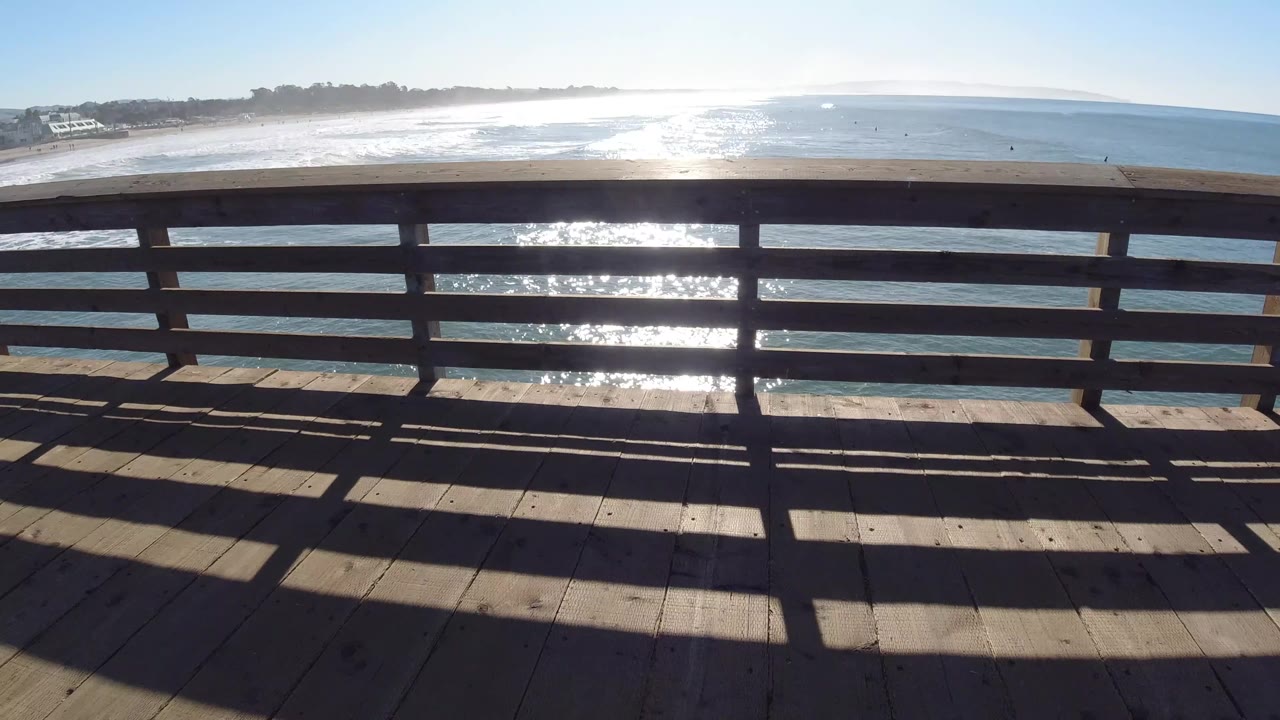 Pismo Beach water and shoreline sit beyond a wooden railing, with bright reflection near the center and long railing shadows stretching across the deck, with no visible people.