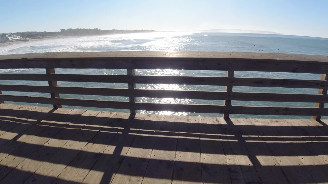 Pismo Beach surf and blue water are seen through horizontal wooden rails, with a bright band of sun reflection below the horizon and no visible people.