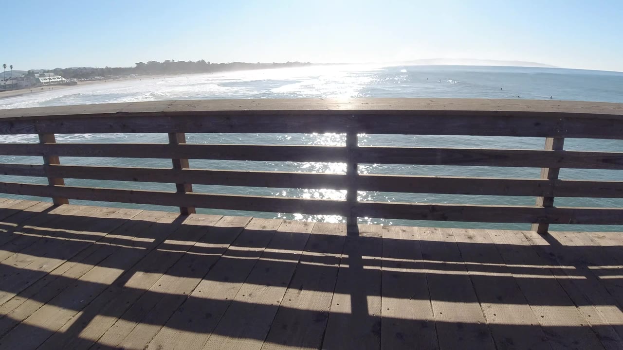 Pismo Beach shoreline and ocean appear behind a dark wooden railing, while the deck foreground is patterned by rail shadows and no visible people appear in the frame.