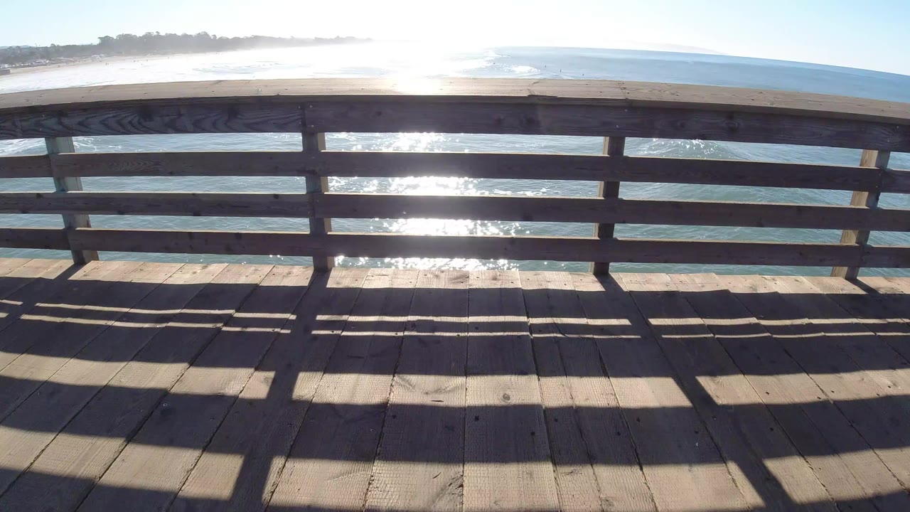 At Pismo Beach, California, this frame holds the wooden deck and rail in the foreground while sunlight flashes through the railing onto the ocean, with no visible people.