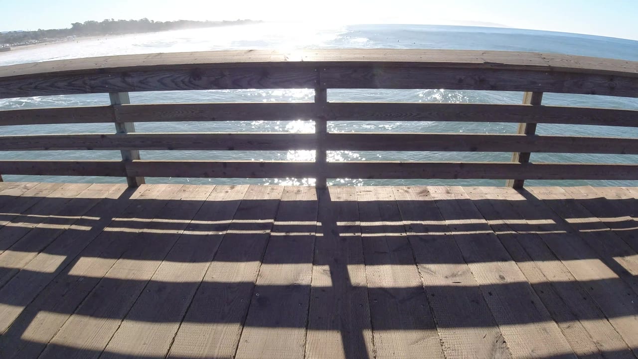 At Pismo Beach, California, the pier railing crosses the middle of the frame with blue water, sandy shore, and bright reflected light behind it, with no visible people.