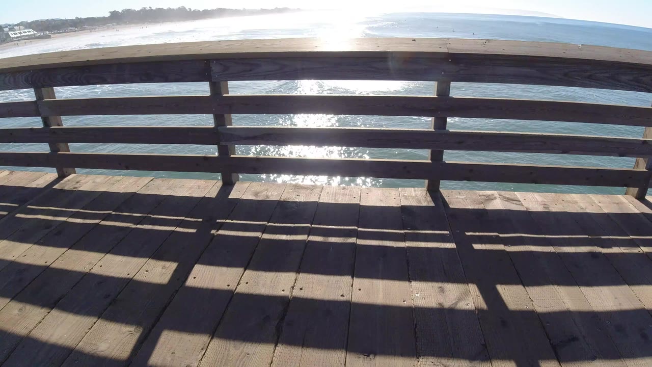Wooden bridge deck and railing in Pismo Beach, California, with bright sun glare on the ocean and tiny distant people on the sandy shoreline.