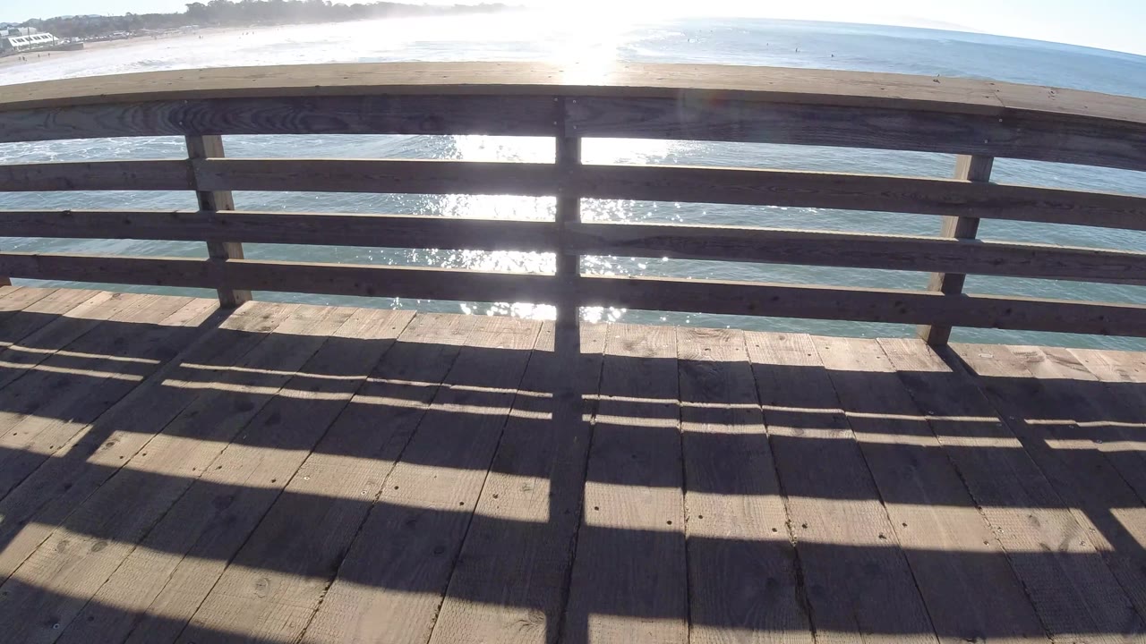 A central railing post divides the Pismo Beach view, with sparkling ocean water behind it and long shadow bands across the bridge deck.