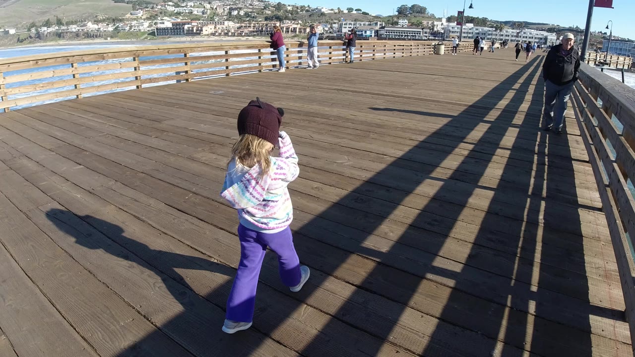 A young girl in a purple hat walks away from the camera on a wide wooden pier in Pismo Beach, with adults ahead, railings, water, and hillside buildings in view.